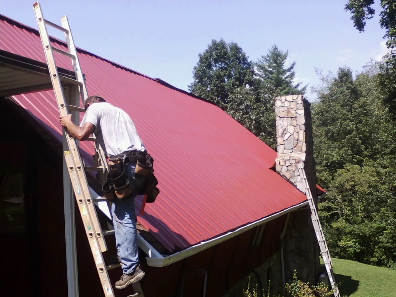 Roofer on a ladder beside a red metal roof and stone chimney — Soffit & Fascia Repair by Roofing Brothers in Lothian